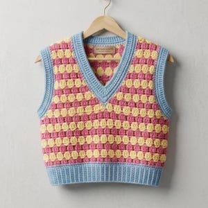 May include: A handmade, colorful crochet vest in a bubble stitch pattern. The vest features alternating rows of pink and yellow, with light blue trim around the neckline, armholes, and hem. The label reads "ARTISAN KNITS".