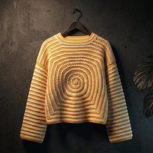 Może przedstawiać: Sweter z długim rękawem wykonany na szydełku, z kolistym, promienistym wzorem w naprzemienne paski w kolorze musztardowym i kremowobiałym. Odzież posiada okrągły dekolt i prosty dół, zawieszona na czarnym wieszaku.