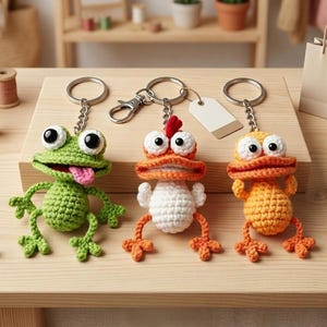 Op de afbeelding: Drie handgemaakte gehaakte sleutelhangers met een groene kikker met een roze tong, een wit en oranje kip met een rode kam, en een oranje eend. Elk figuurtje heeft grote witte ogen met zwarte pupillen en is bevestigd aan een metalen ring.