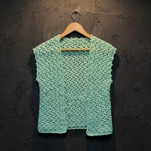 Può includere: Un gilet senza maniche all'uncinetto color verde menta con un design aperto sul davanti e un motivo a punti strutturati ripetuti. Il capo è esposto su una gruccia di legno, mettendo in risalto l'intricata lavorazione del filato leggero e il colore verde chiaro uniforme.