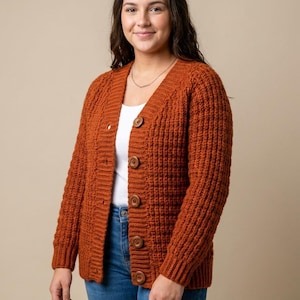 Peut inclure: Un cardigan en tricot orange brûlé à manches longues avec un motif gaufré texturé, une encolure en V et cinq gros boutons ronds marron sur le devant. Le cardigan est porté sur un haut blanc uni et un jean bleu en denim.