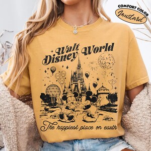 May include: Mustard-colored t-shirt with a black graphic of the Walt Disney World castle, Mickey and Minnie Mouse, and the text "Walt Disney World" and "The happiest place on earth". The shirt has a comfort color and a vintage design.