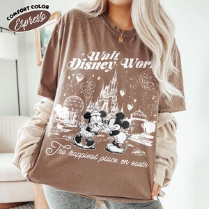 May include: Espresso-colored t-shirt with a graphic of Mickey and Minnie Mouse in front of Cinderella's castle. The text "Walt Disney World" and "The happiest place on earth" are printed on the shirt. The shirt is a Comfort Color brand.