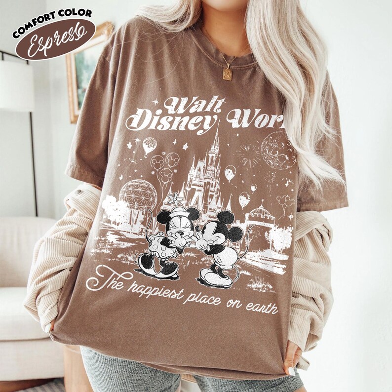May include: Espresso-colored t-shirt with a graphic of Mickey and Minnie Mouse in front of Cinderella's castle. The text "Walt Disney World" and "The happiest place on earth" are printed on the shirt. The shirt is a Comfort Color brand.