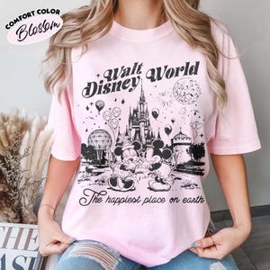 May include: Pink Comfort Color Blossom t-shirt with a black graphic of the Walt Disney World logo, Cinderella's castle, Mickey and Minnie Mouse, and the phrase "The happiest place on earth."