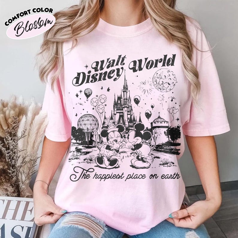 May include: Pink Comfort Color Blossom t-shirt with a black graphic of the Walt Disney World logo, Cinderella's castle, Mickey and Minnie Mouse, and the phrase "The happiest place on earth."