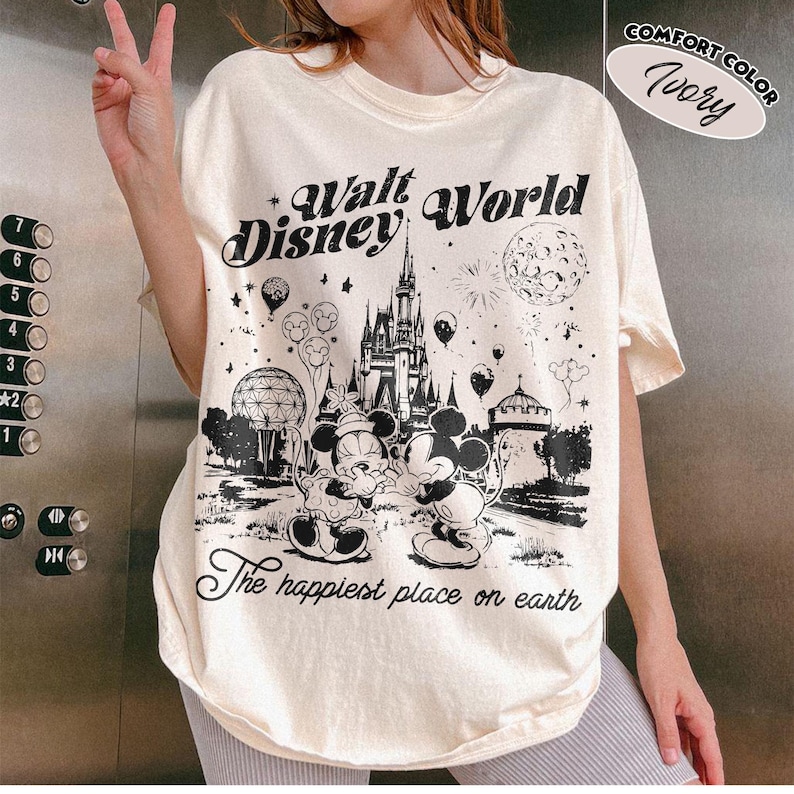 May include: Ivory-colored Walt Disney World graphic tee featuring Mickey and Minnie Mouse, a castle, and the phrase "The happiest place on earth." The shirt has a comfort color and a vintage design.