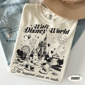 May include: Ivory-colored Comfort Colors t-shirt featuring a black graphic of Mickey and Minnie Mouse in front of Cinderella's castle at Walt Disney World. The shirt also has the text "Walt Disney World" and "The happiest place on earth."