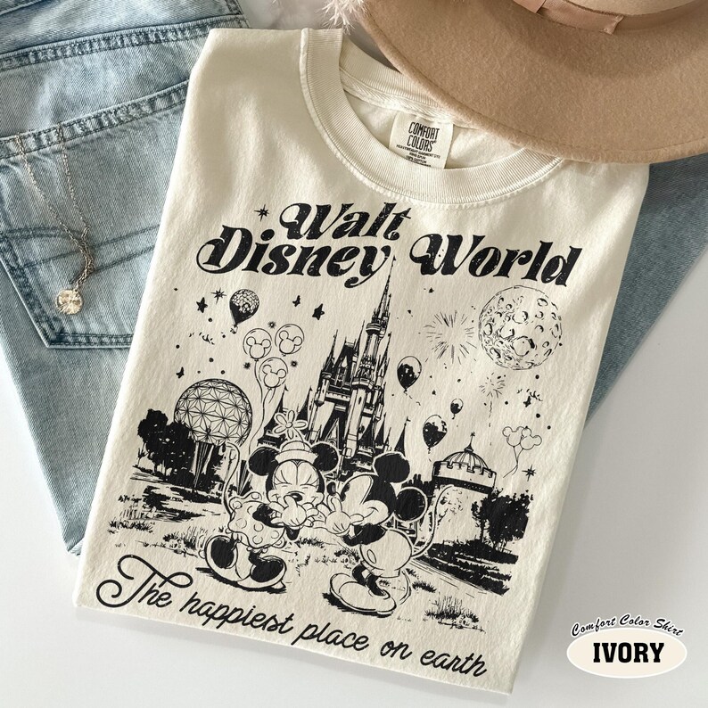 May include: Ivory-colored Comfort Colors t-shirt featuring a black graphic of Mickey and Minnie Mouse in front of Cinderella's castle at Walt Disney World. The shirt also has the text "Walt Disney World" and "The happiest place on earth."