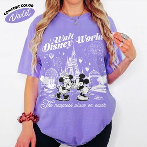 May include: A violet Comfort Colors t-shirt featuring a graphic of Mickey and Minnie Mouse, the text "Walt Disney World", and "The happiest place on earth". The shirt has short sleeves and a crew neck.