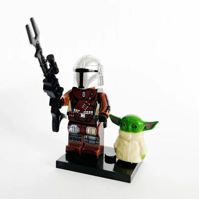 May include: A Lego Mandalorian figure with a silver helmet, brown armor, and a black weapon. Beside it is a green-skinned, large-eared Grogu figure in a tan robe. Both figures stand on a black base.