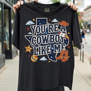 May include: A black short-sleeve t-shirt featuring a graphic of the state of Texas filled with a starry night sky pattern. The text reads "YOU'RE A COWBOY LIKE ME" in white letters, surrounded by illustrations of a cowboy hat, boots, a sheriff badge, a rocket, and a ball.