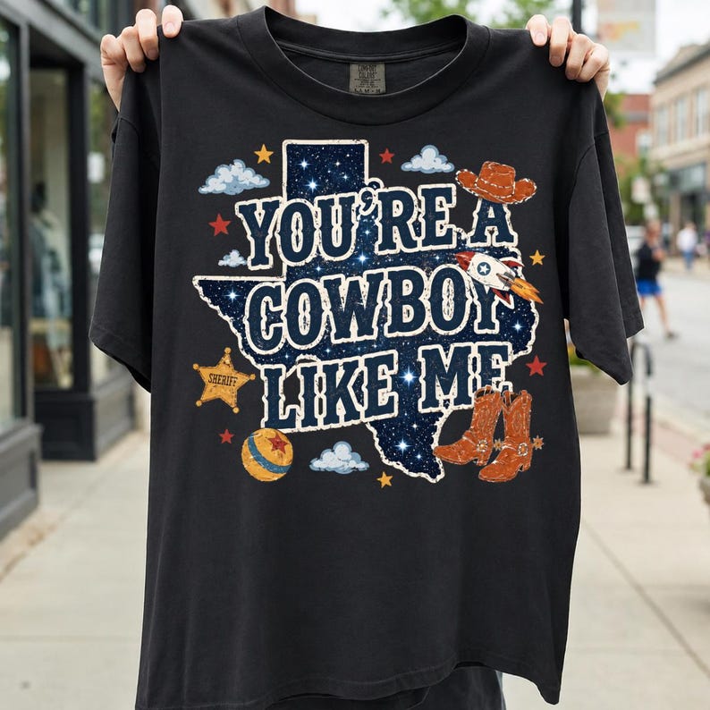 May include: A black short-sleeve t-shirt featuring a graphic of the state of Texas filled with a starry night sky pattern. The text reads "YOU'RE A COWBOY LIKE ME" in white letters, surrounded by illustrations of a cowboy hat, boots, a sheriff badge, a rocket, and a ball.