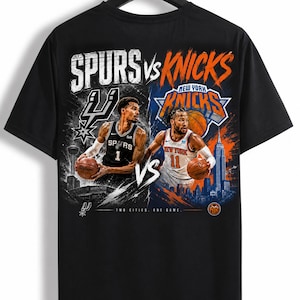 May include: A black t-shirt featuring a graphic print of a basketball game between the Spurs and the Knicks. The design includes two players, team logos, city skylines, and the text "SPURS VS KNICKS" and "TWO CITIES. ONE GAME." in a distressed style.