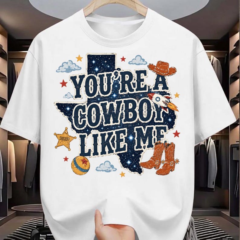 May include: A white short-sleeved t-shirt featuring a graphic of the state of Texas filled with a starry night sky pattern. The text reads YOU'RE A COWBOY LIKE ME, surrounded by illustrations of a cowboy hat, rocket, sheriff star, and boots.