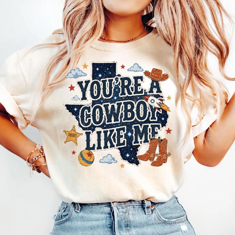 May include: A cream-colored t-shirt featuring a dark blue graphic of the state of Texas filled with a starry night pattern. The text reads "YOU'RE A COWBOY LIKE ME" alongside illustrations of a cowboy hat, a rocket, a sheriff star, and boots.