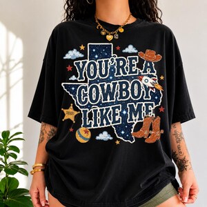 May include: A black short-sleeve t-shirt featuring a graphic of the state of Texas filled with a starry night sky pattern. The text reads "YOU'RE A COWBOY LIKE ME" in white letters, surrounded by illustrations of a cowboy hat, rocket, sheriff badge, and boots.