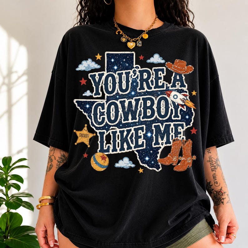 May include: A black short-sleeve t-shirt featuring a graphic of the state of Texas filled with a starry night sky pattern. The text reads "YOU'RE A COWBOY LIKE ME" in white letters, surrounded by illustrations of a cowboy hat, rocket, sheriff badge, and boots.