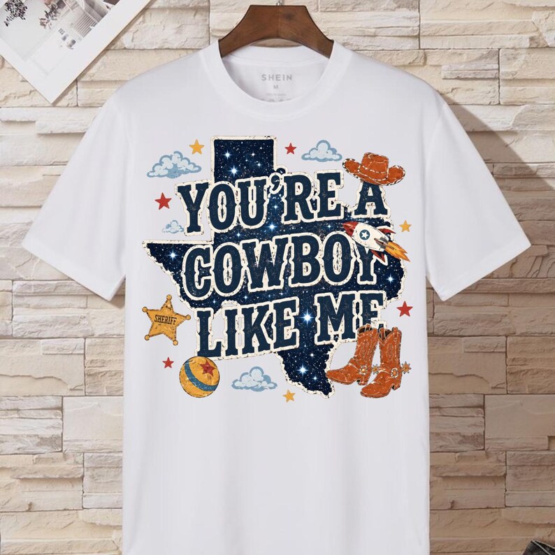 May include: A white short-sleeved t-shirt featuring a graphic of the state of Texas filled with a dark blue starry night pattern. The text reads YOU'RE A COWBOY LIKE ME, surrounded by illustrations of a cowboy hat, rocket, sheriff star, and boots.