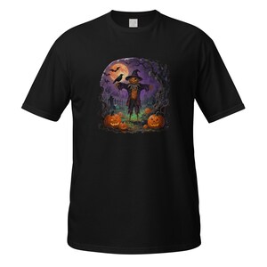 Pode incluir: Uma t-shirt preta de manga curta com uma estampa gráfica de um espantalho com cabeça de abóbora e chapéu, num campo com abóboras esculpidas, um corvo preto, uma lua cheia grande e árvores nuas sob um céu de tom roxo escuro.