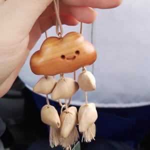 May include: A wooden cloud-shaped ornament with a smiling face, hanging from a beige string. Below the cloud are several teardrop-shaped wooden pieces with tassels. The ornament is held up by a hand.