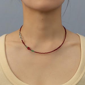 May include: A delicate necklace featuring small, dark red beads. The necklace has a silver clasp and a section with a larger red bead and two small green beads. The necklace is worn around the neck.