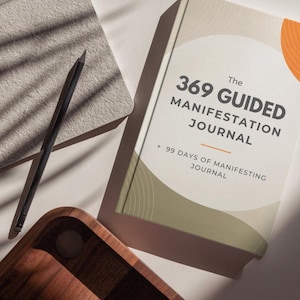 May include: A hardback book titled The 369 Guided Manifestation Journal + 99 Days of Manifesting Journal with a cream cover, olive green circular graphic, and orange accent. A dark pencil rests on a textured grey notepad next to the book.