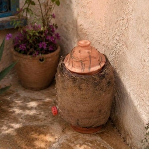 Peut inclure: Une jarre à eau en terre cuite avec un couvercle, enveloppée dans un matériau semblable à du jute. La jarre a un robinet rouge et repose sur une surface en pierre. Une plante en pot avec des fleurs violettes est en arrière-plan.