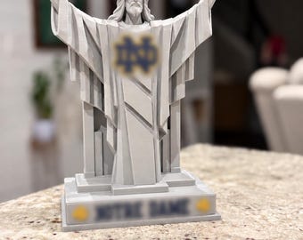 May include: A light grey 3D-printed statue of a figure with arms outstretched, wearing long robes. The figure features a blurred logo on the chest and a rectangular base with the blurred text "NOTRE DAME" flanked by two small yellow circular icons.