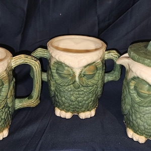 May include: A set of three ceramic owl-shaped vessels in shades of green and cream. Two vessels feature single handles, while the central vessel has two side handles. The third vessel on the right includes a matching circular lid with a small handle.