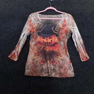 May include: A long-sleeve shirt featuring a central graphic of a jack-o'-lantern wearing a black hat, surrounded by orange and brown autumn leaves, swirling filigree patterns, and a spiderweb design on a beige and brown mottled background.