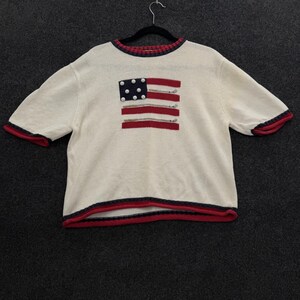 May include: A short-sleeved cream-colored knit sweater featuring a central graphic of a flag design composed of red stripes and a blue square with white circular accents. The neckline, sleeve cuffs, and bottom hem are trimmed in red and navy blue.
