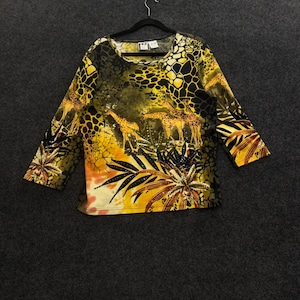 Könnte beinhalten: Ein langärmeliges Oberteil mit einem lebendigen Giraffenmuster in Gelb, Orange und Schwarz. Das Design zeigt Illustrationen von Giraffen inmitten von tropischem Blattwerk und großen Blumen, mit einem Rundhalsausschnitt und geradem Saum.