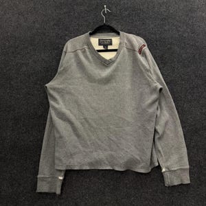 May include: A grey long-sleeve V-neck jumper with a cream-coloured inner lining at the neckline. The garment features the Abercrombie & Fitch brand label inside the collar and a small red embroidered detail on the left shoulder area.
