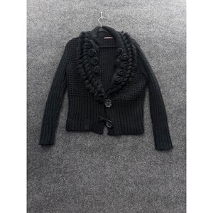 May include: A black knit cardigan featuring a wide shawl collar adorned with decorative fabric rosettes. The garment has a textured waffle-knit upper section, a ribbed lower hem, long sleeves, and two large, dark, flower-shaped buttons at the front.