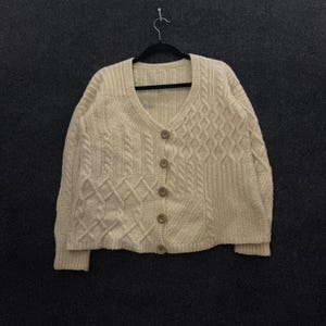 Pode incluir: Um cardigã de cor creme com decote em V, apresentando um mosaico de vários padrões de malha trançada. A peça possui mangas compridas, uma bainha canelada e é fechada na frente com cinco botões redondos de cor castanho-claro. A textura é visível.