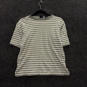 Peut inclure: Un t-shirt à manches courtes avec un motif à rayures horizontales vert sauge et blanc cassé. Le vêtement présente une encolure ronde, un ourlet droit avec de petites fentes latérales, et est présenté sur un cintre noir sur fond gris foncé.