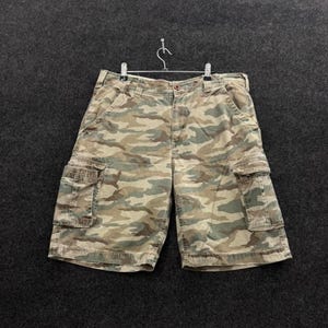 May include: Camouflage cargo shorts in shades of green, brown, and tan. The shorts have two side cargo pockets and are hanging on a metal hanger. The shorts are made of a durable fabric.