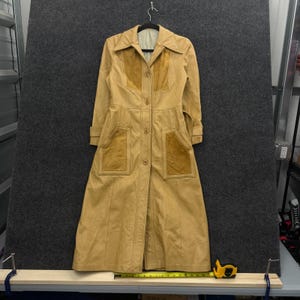 Peut inclure: Un long manteau en cuir beige doté d'un col pointu, d'une fermeture boutonnée sur le devant et de deux grandes poches plaquées. Le manteau présente des empiècements en daim contrastants sur la poitrine et les poches. Un mètre ruban est visible.