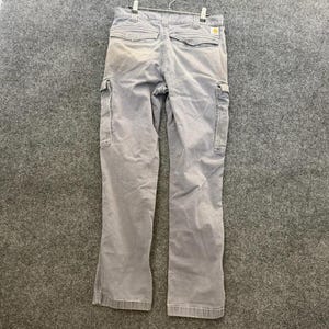 以下が含まれることがあります： 複数のポケットとCarharttのロゴパッチが付いた、明るいグレーのカーゴパンツ。パンツは、グレーの背景に金属製のハンガーに吊るされています。パンツは色あせた外観をしています。