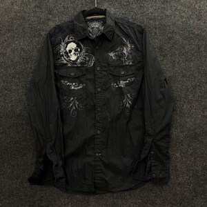 Puede incluir: Una camisa negra de manga larga con botones, un gráfico de calavera blanca en el pecho izquierdo, bordados grises en forma de volutas y dos bolsillos con solapa. El texto "Born to Renegade" está bordado debajo del bolsillo izquierdo.