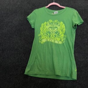 May include: A bright green short-sleeved t-shirt featuring a large, lime green graphic print on the chest. The design consists of an ornate, symmetrical emblem with leafy flourishes and a central heart-like shape. The shirt has a crew neckline.