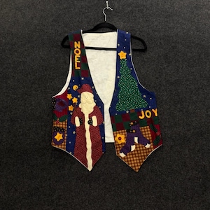 May include: A sleeveless waistcoat featuring a festive holiday theme with a white background and colourful fabric appliques. The design includes a Santa Claus figure, a Christmas tree, stars, and the words NOEL and JOY in yellow block letters.