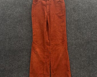 May include: A pair of rust-colored corduroy pants featuring a high-waisted design, a button and zipper fly closure, two front pockets, and a straight-leg cut. The pants are suspended from a metal hanger, showcasing the textured fabric and stitching.