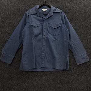 May include: A long-sleeved, blue button-down shirt with two chest pockets. The shirt has a wide collar and buttoned cuffs. The garment is hanging on a black hanger against a dark gray background.