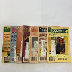 May include: A collection of seven vintage Workbasket and Home Arts magazines arranged side-by-side. Each issue features a distinct colour cover, including yellow, tan, brown, and blue, with bold typography and various craft project illustrations.