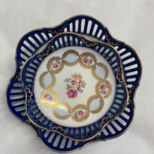 May include: A set of two porcelain plates with a deep blue reticulated border and gold trim. The centre features a white background with a floral bouquet and a circular wreath pattern of gold leaves interspersed with small pink rose clusters.