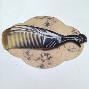 May include: A dark brown and translucent horn comb carved into the shape of a rooster. The comb features a series of fine teeth on one side and a detailed bird head with a small yellow eye on the other, resting on a beige oval fabric base.