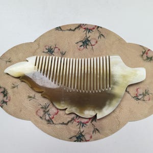 May include: A handcrafted comb with a unique shape, featuring a blend of light and dark tones. The comb is set on a decorative, floral-patterned background, showcasing a blend of natural materials and artistic design.