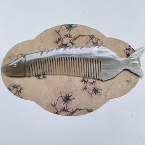 May include: A fish-shaped comb with a pearlescent finish. The comb has a series of teeth and a tail fin. The fish comb is set on a beige, cloud-shaped background with a floral pattern.